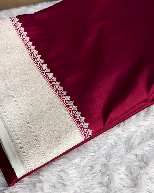 A folded deep maroon silk saree with a wide silver border and intricate geometric embroidery, displayed on a white faux fur surface with a cardboard box in the background.