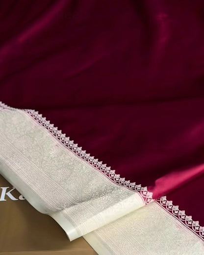 A close-up of a deep magenta silk saree with a wide cream-colored border featuring intricate silver zari embroidery, draped over a brown box labeled 'kaashivastra' on a white textured rug.