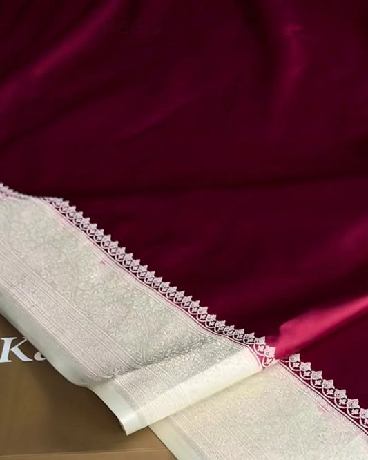 A close-up of a deep magenta silk saree with a wide cream-colored border featuring intricate silver zari embroidery, draped over a brown box labeled 'kaashivastra' on a white textured rug.