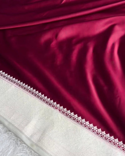 A close-up of a deep magenta satin fabric draped elegantly, showing its smooth texture and a decorative white border with a geometric pattern, laid over a white shag rug.
