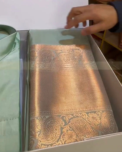 A folded metallic gold and silver paisley patterned fabric, likely a brocade sash or scarf, displayed inside a white box next to a light green shirt. A hand is visible pointing towards the top of the fabric.
