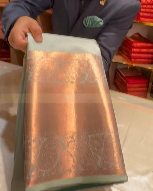 A person in a navy blazer holds up a folded metallic fabric, likely a silk saree, showing its sheen and woven pattern. The fabric features a prominent coppery-gold central panel with a pale sage green border.