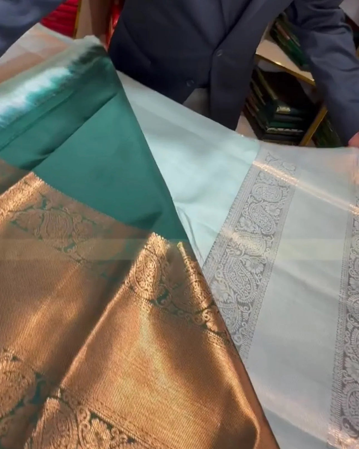 A close-up of a folded and draped silk saree, showing a teal green body, a wide metallic gold zari border, and a pale blue-green border with intricate woven patterns. A person in a dark blazer is partially visible in the background.
